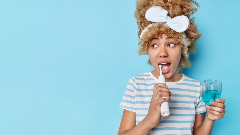 photo-blonde-curly-woman-brushes-teeth-with-electric-brush-holds-glass-mouthwash-fresh-breath-isolated-blue-background-blank-space-your-promotional-content-dental-care-concept