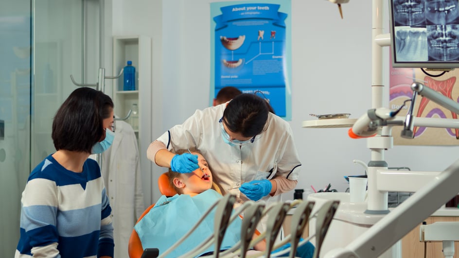 stomatologist-talking-with-mother-kids-lighting-lamp-examining-little-girl-standing-near-stomatological-chair-pediatric-dentist-speaking-woman-while-nurse-preparing-sterilized-tools